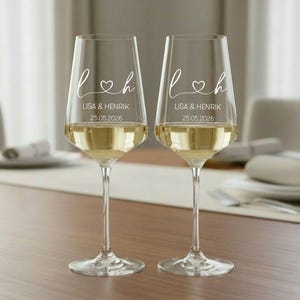 May include: Two clear glass wine glasses with long stems, each featuring white printed text that reads L and H separated by a heart symbol, followed by the names LISA & HENRIK and the date 25.05.2026. The glasses are filled with a pale liquid.