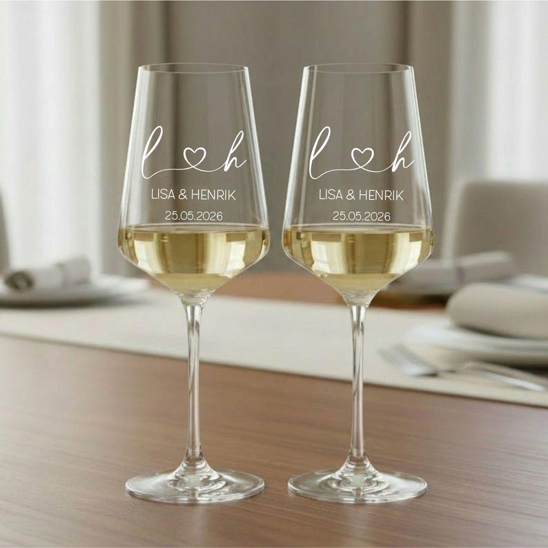 May include: Two clear glass wine glasses with long stems, each featuring white printed text that reads L and H separated by a heart symbol, followed by the names LISA & HENRIK and the date 25.05.2026. The glasses are filled with a pale liquid.