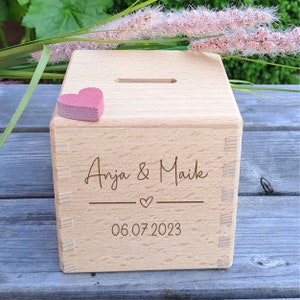 Könnte beinhalten: Eine Holzkiste mit einem Schlitz oben zum Geld sparen. Die Kiste ist mit den Namen "Anja & Maik" und dem Datum "06.07.2023" graviert. Ein kleines rosa Herz liegt oben auf der Kiste.