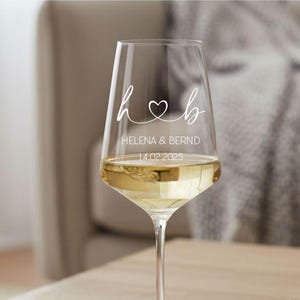 May include: A clear glass wine glass featuring white printed text and graphics. The design includes the lowercase letters h and b connected by a heart symbol, followed by the names HELENA & BERND and the date 14.02.2025 in a clean font.