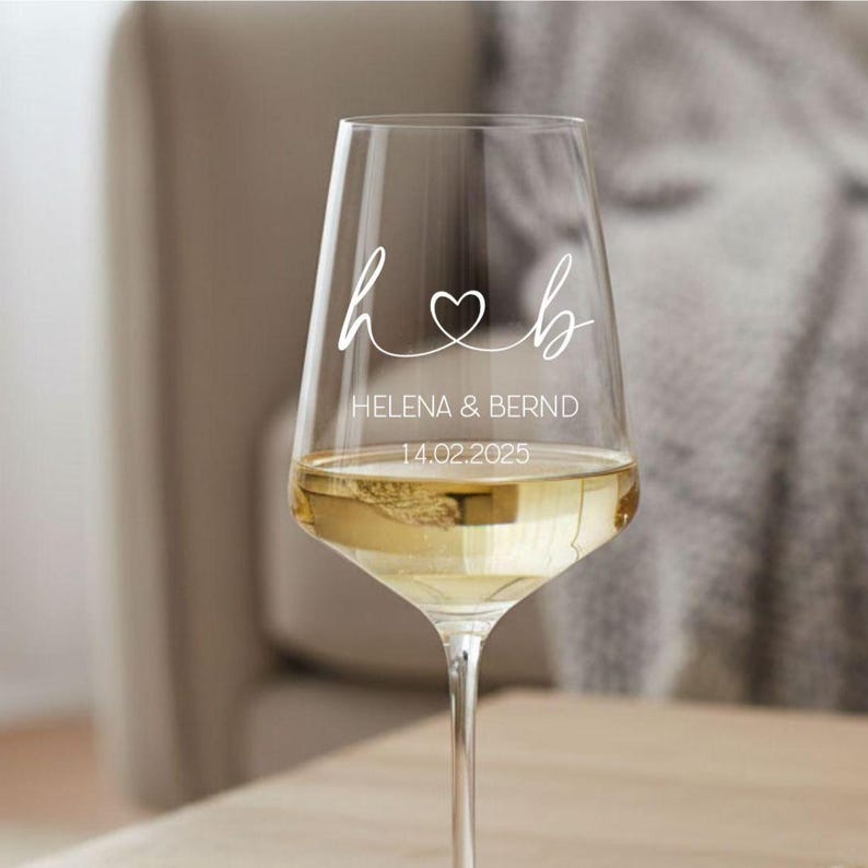 May include: A clear glass wine glass featuring white printed text and graphics. The design includes the lowercase letters h and b connected by a heart symbol, followed by the names HELENA & BERND and the date 14.02.2025 in a clean font.
