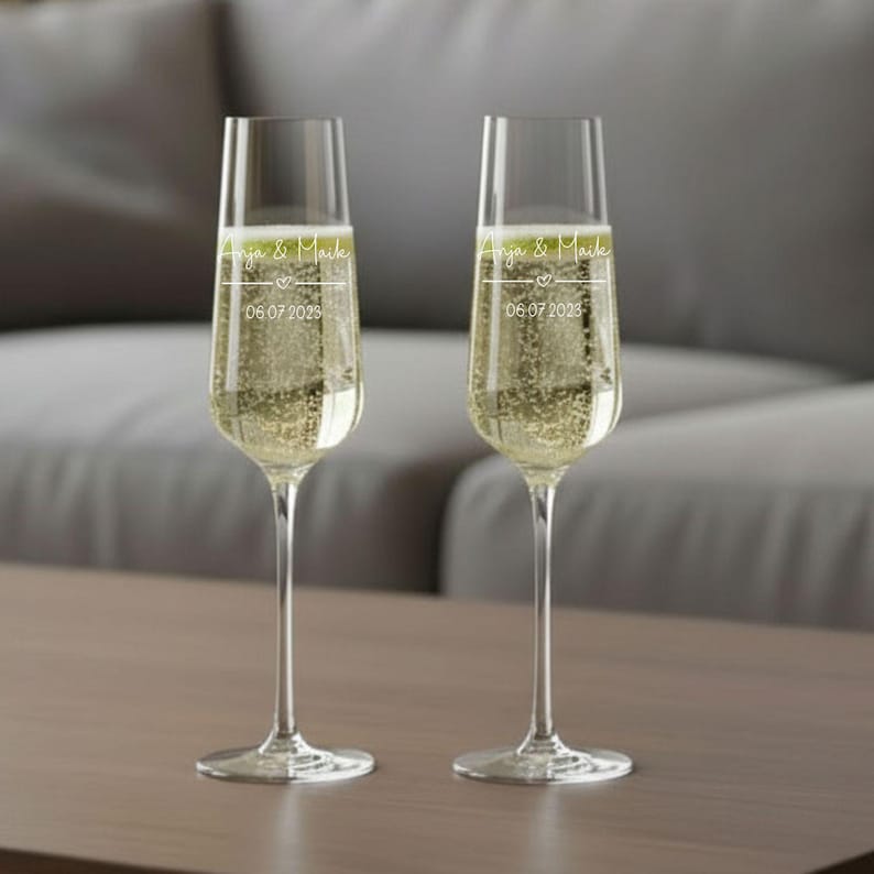 Puede incluir: Dos copas de champ&aacute;n llenas de l&iacute;quido espumoso, probablemente champ&aacute;n o vino espumoso, sobre una superficie de madera. Las copas tienen el texto personalizado "Anja & Maik" y la fecha "06.07.2023". El fondo est&aacute; borroso, sugiriendo un interior.