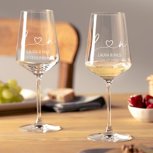 May include: Two clear glass wine glasses featuring white etched text. Each glass displays the letters L and N connected by a heart symbol, followed by the names LAURA & NILS and the date 15.03.2025 printed in a clean, sans-serif font.