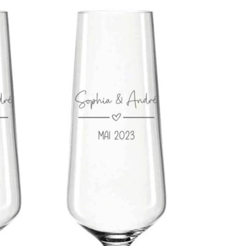 Puede incluir: Una flauta de champ&aacute;n transparente con los nombres "Sophia & Andre" en escritura elegante, un peque&ntilde;o coraz&oacute;n y la fecha "MAI 2023" impresa debajo. La cristaler&iacute;a est&aacute; dise&ntilde;ada para una boda o evento especial.