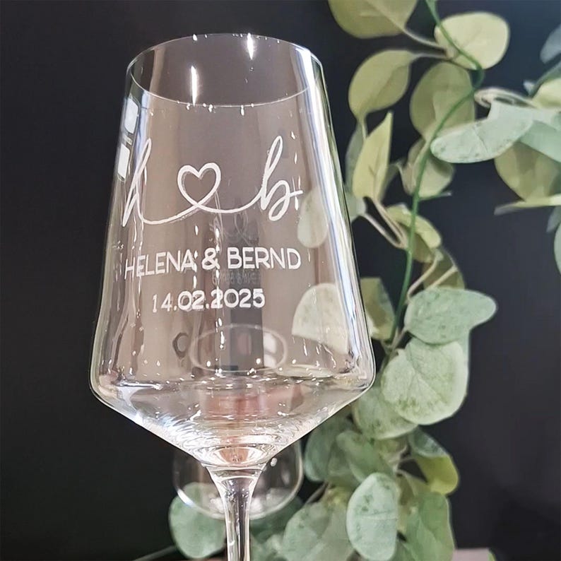 May include: A clear glass wine glass featuring white etched text and graphics. The design includes a stylized heart connecting the letters h and b, followed by the names HELENA & BERND and the date 14.02.2025 printed in a clean font.