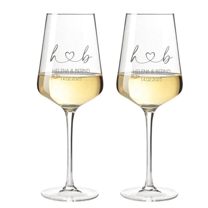 May include: Two clear glass wine glasses filled with white wine. Each glass features a black printed design with the letters h and b connected by a heart, the names HELENA & BERND, and the date 14.02.2025 printed in a clean, sans-serif font.