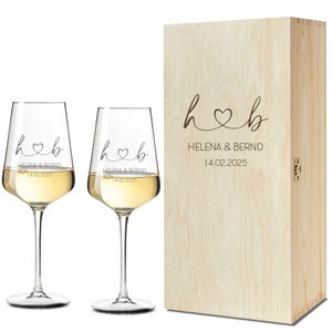 Könnte beinhalten: Zwei klare Weingläser gefüllt mit Weißwein, neben einer Holzkiste. Die Gläser und die Kiste tragen die Initialen "h♡b" und den Text "HELENA & BERND 14.02.2025". Die Kiste hat einen Klappdeckel und einen Metallverschluss.