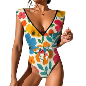 Op de afbeelding: Badpak uit één stuk met een bloemenpatroon. Het heeft een diepe V-hals, zwarte bies en een strikceintuur in de taille. Het bloemmotief omvat oranje, gele, blauwe en rode bloemen met groene bladeren op een gebroken witte achtergrond.