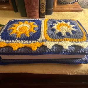 May include: A crocheted book cover with a blue, white and yellow design featuring two suns. The cover is made with a variety of stitches and textures.