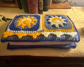 May include: A crocheted book cover with a blue, white, and yellow design featuring two suns. The cover is made with a variety of stitches and textures.