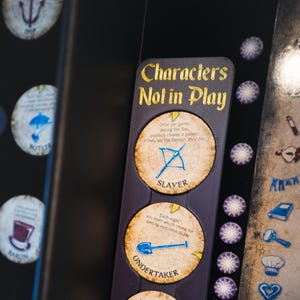 May include: A close-up of a board game component labeled "Characters Not in Play." It features three character cards: Slayer, Undertaker, and Recluse. Each card has a unique icon and description. The background is dark with other game elements.