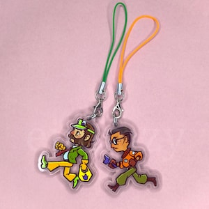 May include: Two acrylic charms featuring cartoon characters. One character has brown hair, a green jacket, yellow trousers, and a green cap. The other character has dark hair, glasses, an orange jacket, and green trousers. Both have colourful lanyards.