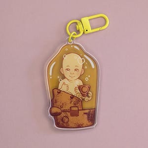 Könnte beinhalten: Ein gelber Schlüsselanhänger mit einem durchsichtigen Plastik-Anhänger, der ein Cartoon-Baby in einem gelben Overall zeigt, das einen Daumen nach oben zeigt. Das Baby befindet sich in einem durchsichtigen Plastikbehälter mit braunem und goldenem Design.
