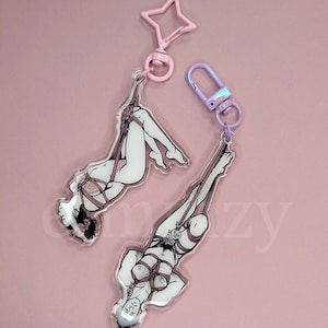 Puede incluir: Dos llaveros acrílicos con ilustraciones de figuras en poses de bondage. Las figuras están representadas en blanco y negro con detalles de cuerda rosa. Cada llavero tiene un cierre de diferente color, uno rosa en forma de estrella y otro iridiscente.