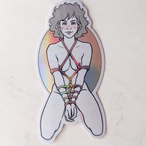 Puede incluir: Una pegatina con un dibujo lineal de una persona con el pelo rizado, atada con cuerdas de colores, sobre un fondo holográfico.