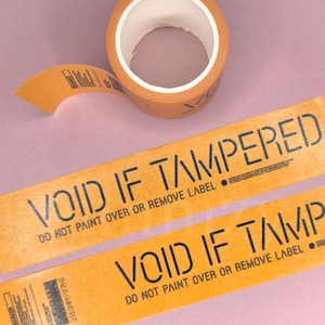 Può includere: Un rotolo di nastro adesivo arancione con testo nero che recita VOID IF TAMPERED e DO NOT PAINT OVER OR REMOVE LABEL. Il nastro è mostrato come rotolo e come due strisce piatte, con un codice a barre e ulteriori piccole istruzioni stampate.