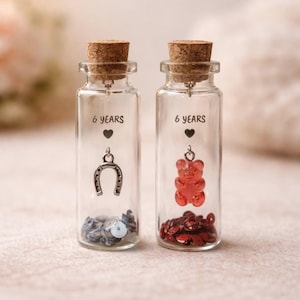May include: Two small glass bottles with cork stoppers. One bottle contains a silver horseshoe charm and silver sequins, the other a red gummy bear charm and red sequins. Both bottles have the text "6 years" and a heart.