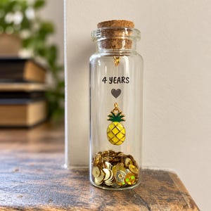 Puede incluir: Una botella de vidrio transparente con tapón de corcho que contiene lentejuelas doradas en la base. Dentro, un dije de piña dorado cuelga bajo un icono de corazón negro y el texto 4 YEARS impreso en la superficie del vidrio.