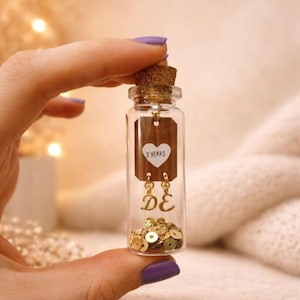 May include: A small glass bottle with a cork stopper, filled with gold sequins. Inside, a wooden tag with "3 YEARS" in a heart shape and gold earrings with "DE" initials. The bottle is held by a hand with purple nail polish.
