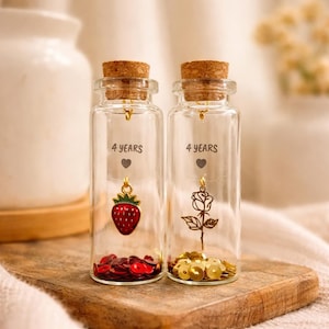 Pode incluir: Duas garrafas de vidro transparente com rolhas de cortiça, cada uma contendo elementos decorativos. Uma garrafa tem um pingente de morango e lantejoulas vermelhas, a outra um pingente de rosa e lantejoulas douradas. Ambas as garrafas estão rotuladas como "4 YEARS".