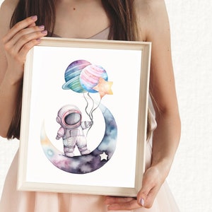 May include: Watercolor art print of an astronaut holding planet and star balloons while standing on a crescent moon. The artwork features a soft color palette of blues, purples, and pinks, perfect for a nursery or children's room.