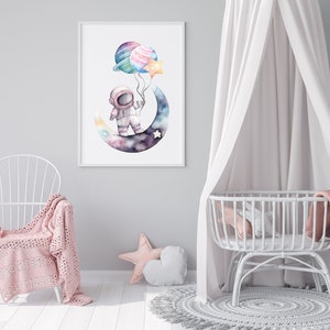 May include: Watercolor print of an astronaut on a crescent moon holding planet and star balloons. The artwork features a soft color palette of blues, purples, and pinks, perfect for a nursery or children's room. The print is framed in a white frame.