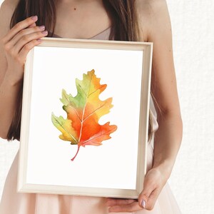 May include: A watercolour painting of a single autumn leaf in shades of yellow, orange, and green. The leaf is framed in a white frame.
