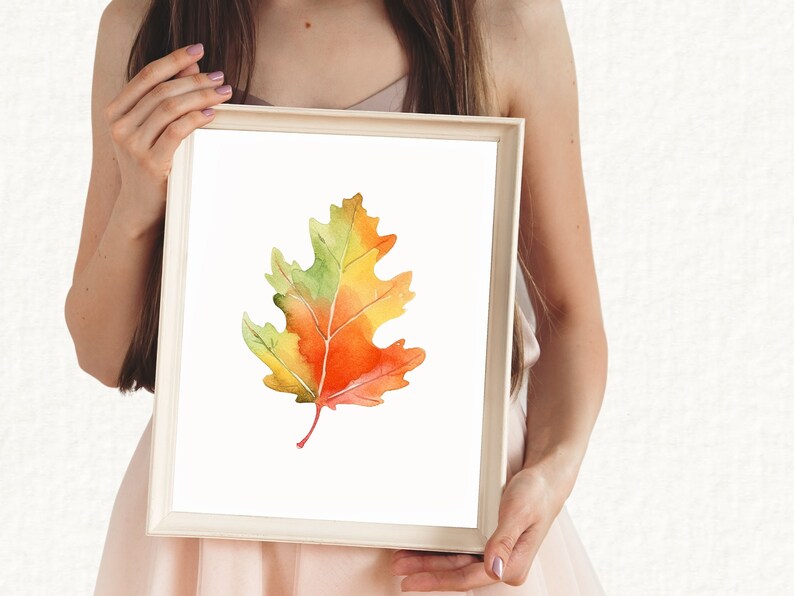 May include: A watercolour painting of a single autumn leaf in shades of yellow, orange, and green. The leaf is framed in a white frame.