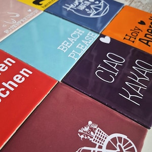 Könnte beinhalten: Eine Sammlung quadratischer Keramikfliesen in verschiedenen Farben, jede mit einem anderen weißen Textdesign. Die Fliesen enthalten Sätze wie "Beach Please" und "Ciao Kakao". Die Fliesen sind zusammen angeordnet und bilden eine farbenfrohe und abwechslungsreiche Darstellung.