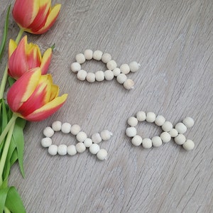 Könnte beinhalten: Drei Holzfischsymbole aus kleinen, runden, hellfarbigen Perlen. Die Fische sind auf einer Holzoberfläche angeordnet, neben einem Strauß roter und gelber Tulpen mit grünen Stielen und Blättern.