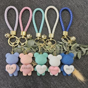 Könnte beinhalten: Fünf gehäkelte Teddybär-Schlüsselanhänger mit herzförmigen Anhängern. Jeder Schlüsselanhänger hat eine andere Farbe und einen personalisierten Namen auf dem herzförmigen Anhänger. Die Schlüsselanhänger sind an geflochtenen Lederbändern mit goldenen Glöckchen befestigt.
