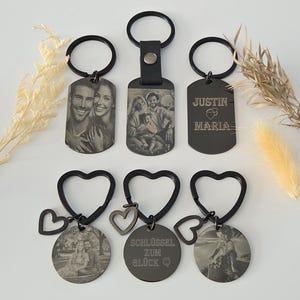 May include: Five different black metal keychains with various designs. One keychain features a rectangular tag with a photo of a couple, another has a rectangular tag with a photo of a family of three, another has a rectangular tag with the text "Justin Maria" and a heart, and the last two keychains have round tags with photos of couples and a heart charm.