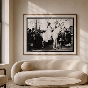 May include: A black and white framed photograph depicting a woman riding a white horse in a parade, with the text "SUFFRAGE PARADE 3/3/13" and "INEZ MILHOLLAND" visible. The print is displayed in a thin wooden frame hanging on a neutral wall.