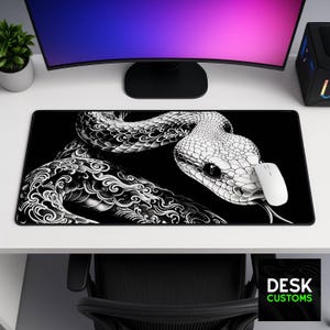 May include: A rectangular desk mat featuring a high-contrast black and white illustration of a snake with intricate scale patterns and swirling designs. A white computer mouse rests on the surface of the mat, which is placed on a white desk.