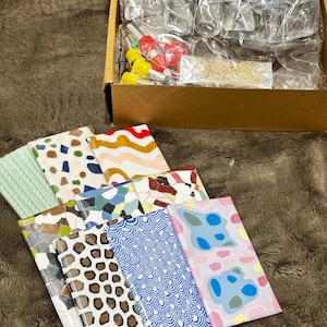 May include: A collection of rectangular clay slabs featuring various patterns including leopard print, blue waves, terrazzo, and abstract shapes, alongside a cardboard box containing metal cookie cutters and small tools in plastic packaging.