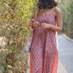 May include: A pink floral print sundress with thin straps. The dress is knee-length and features a repeating pattern of blue floral designs. The person is wearing white sandals and carrying a tote bag with a floral design. A scrunchie is on the wrist.