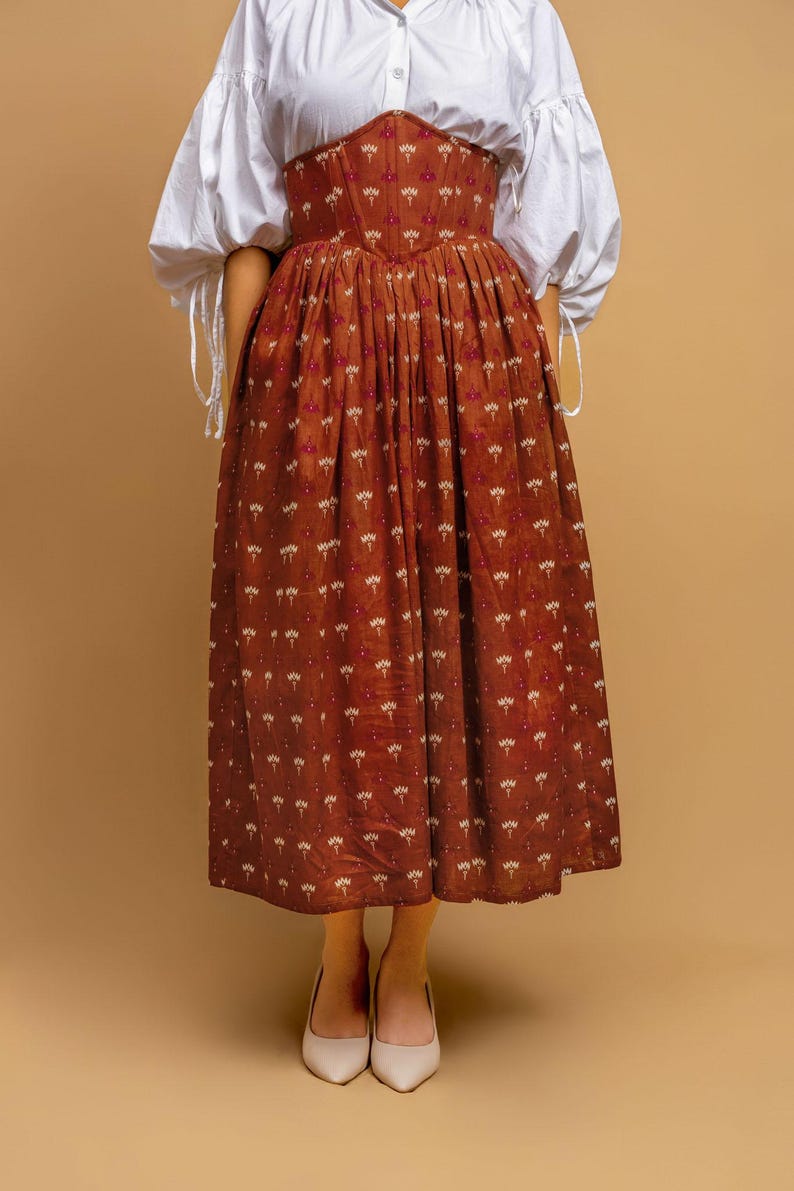 May include: A rust-colored skirt and corset with a white floral pattern, paired with a white blouse with puffy sleeves. The outfit is completed with cream-colored heels, creating a vintage aesthetic.