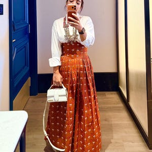 May include: A white button-down shirt paired with a rust-colored, floral-print skirt. The outfit is accessorized with a silver necklace and a white handbag. The person is wearing platform shoes.