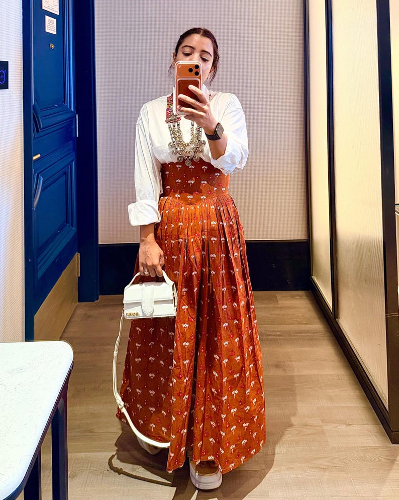 May include: A white button-down shirt paired with a rust-colored, floral-print skirt. The outfit is accessorized with a silver necklace and a white handbag. The person is wearing platform shoes.