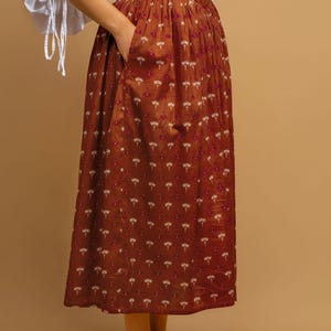 May include: A rust-colored dress with a floral pattern and a white blouse. The dress features a fitted bodice and a flowing skirt with pockets. The white blouse has long sleeves with tie details at the wrists. The outfit is completed with cream-colored heels.