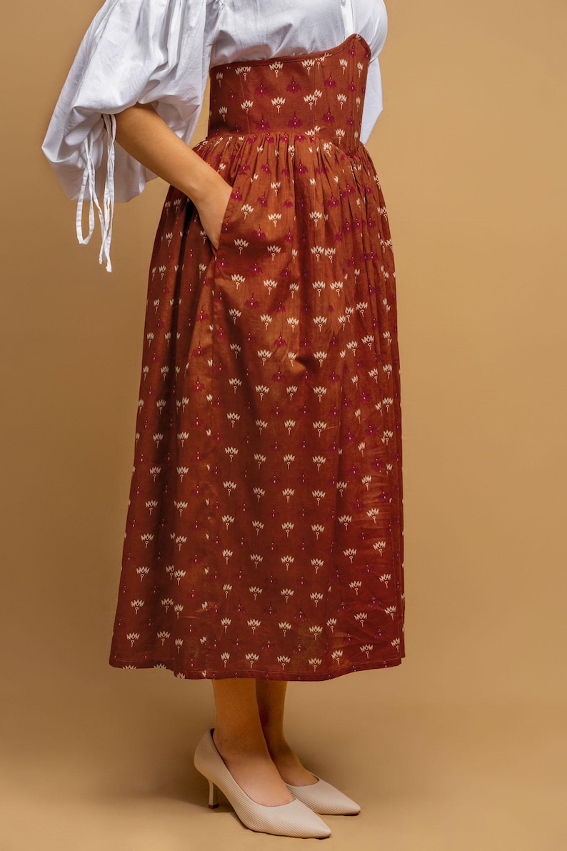 May include: A rust-colored dress with a floral pattern and a white blouse. The dress features a fitted bodice and a flowing skirt with pockets. The white blouse has long sleeves with tie details at the wrists. The outfit is completed with cream-colored heels.