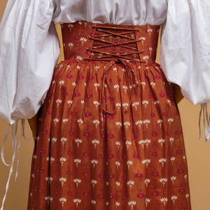 May include: A rust-colored skirt and corset with a floral pattern. The corset has a laced back and the skirt is long and flowing. The outfit is paired with a white blouse with puffy sleeves.