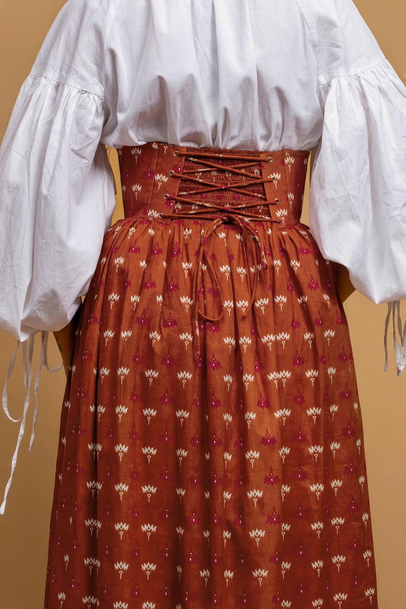 May include: A rust-colored skirt and corset with a floral pattern. The corset has a laced back and the skirt is long and flowing. The outfit is paired with a white blouse with puffy sleeves.