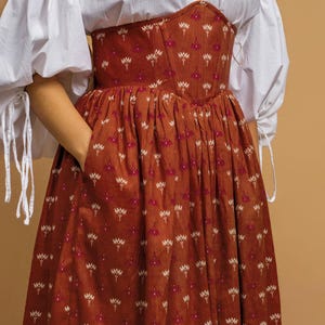 May include: A rust-colored dress with a floral pattern and a fitted bodice. The dress has a full skirt and is paired with a white blouse with puffy sleeves. The floral pattern includes white and pink accents.