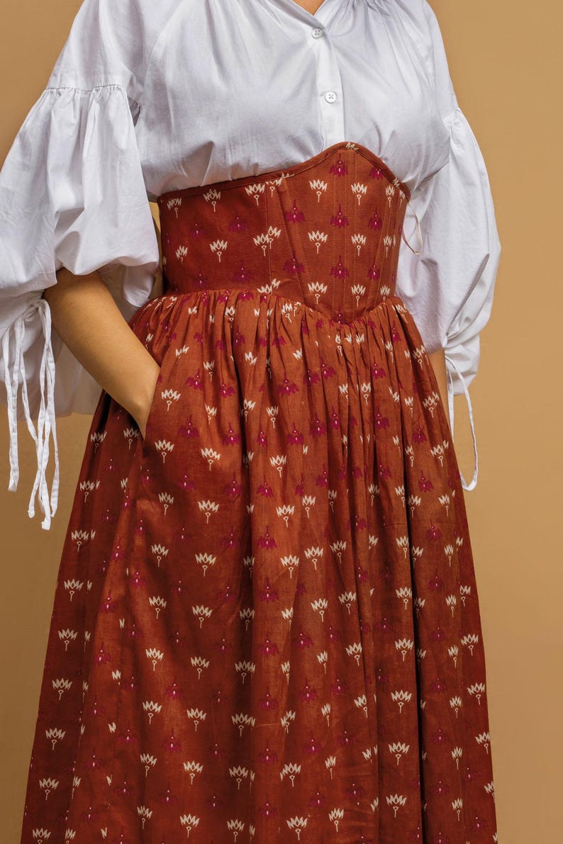 May include: A rust-colored dress with a floral pattern and a fitted bodice. The dress has a full skirt and is paired with a white blouse with puffy sleeves. The floral pattern includes white and pink accents.
