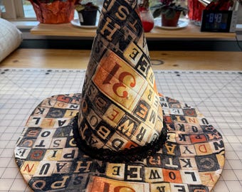May include: A Halloween witch hat with a wide brim and a tall, pointed crown. The hat is decorated with a repeating pattern of letter blocks in orange, black, and cream colors. A black lace trim accents the base of the crown.