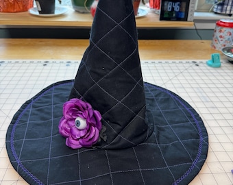 May include: Black quilted witch hat with a wide brim and a pointed top. The hat features a purple flower with an eye in the center. The brim is outlined with purple stitching, and the hat has a diamond pattern.
