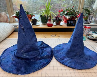 May include: Two navy blue wizard hats with a shimmering, textured fabric. The hats have a wide brim and a tall, pointed crown. The hats are displayed on a light-colored surface.