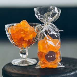 Puede incluir: Un cuenco de vidrio transparente y una bolsa de celofán llena de caramelos de color naranja. Los caramelos tienen un aspecto translúcido. La bolsa tiene una etiqueta marrón con la palabra "ZELT". Los artículos están sobre una superficie de madera oscura.