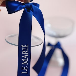 Puede incluir: Cinta de raso azul marino atada en lazo, con el texto "LE MARIÉ" impreso en blanco. La cinta está sujeta a un vaso transparente. El fondo es rosa claro.